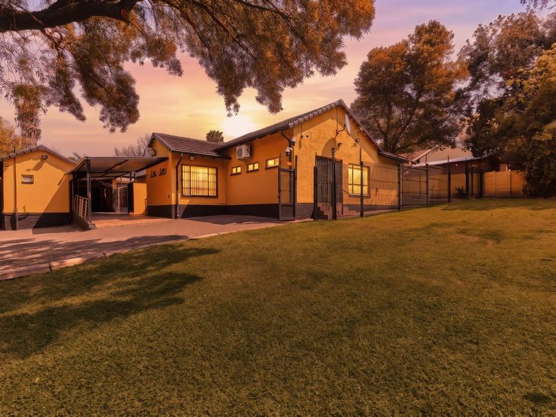 Front view of a yellow house featuring a paved driveway, carport, and secure perimeter fencing set within a landscaped garden.