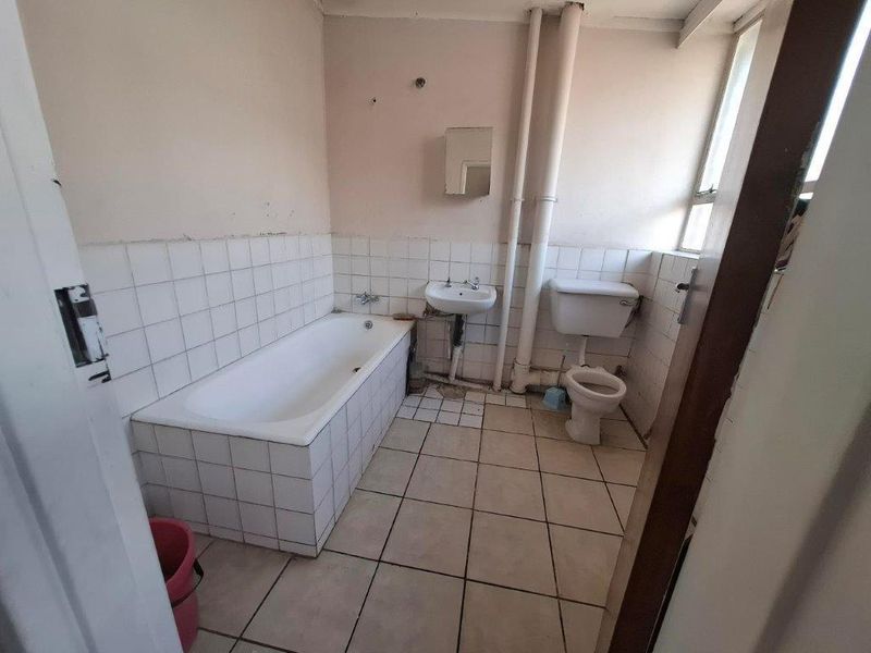 A dated bathroom featuring a tiled bathtub, wall-mounted basin, and toilet, requiring renovation to update the finishes and address exposed plumbing.