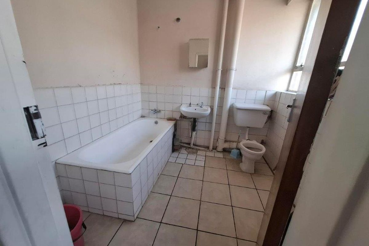 A dated bathroom featuring a tiled bathtub, wall-mounted basin, and toilet, requiring renovation to update the finishes and address exposed plumbing.