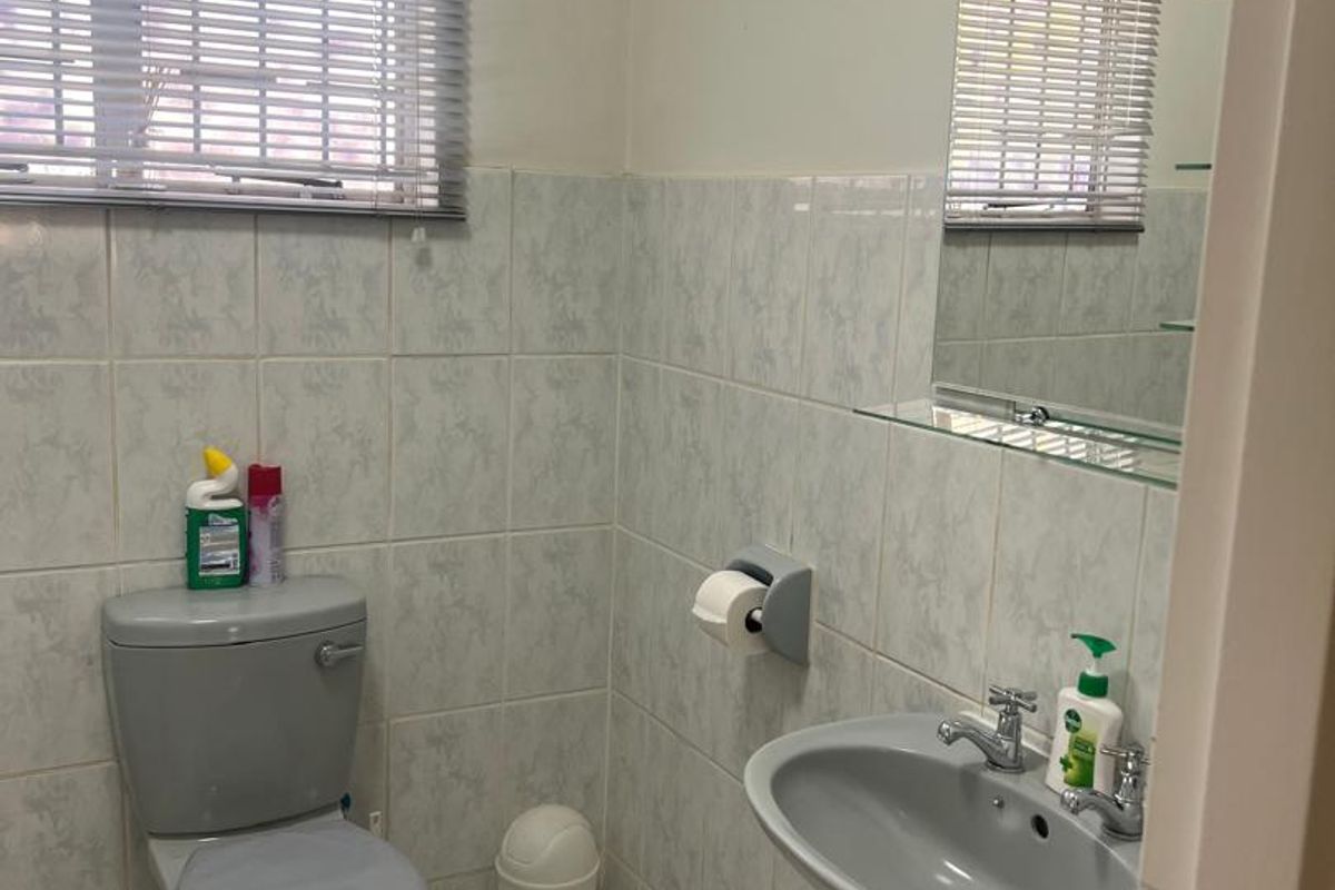 A functional guest bathroom featuring a grey toilet and matching wall-mounted basin, complemented by neutral wall tiles and a window with venetian blinds.