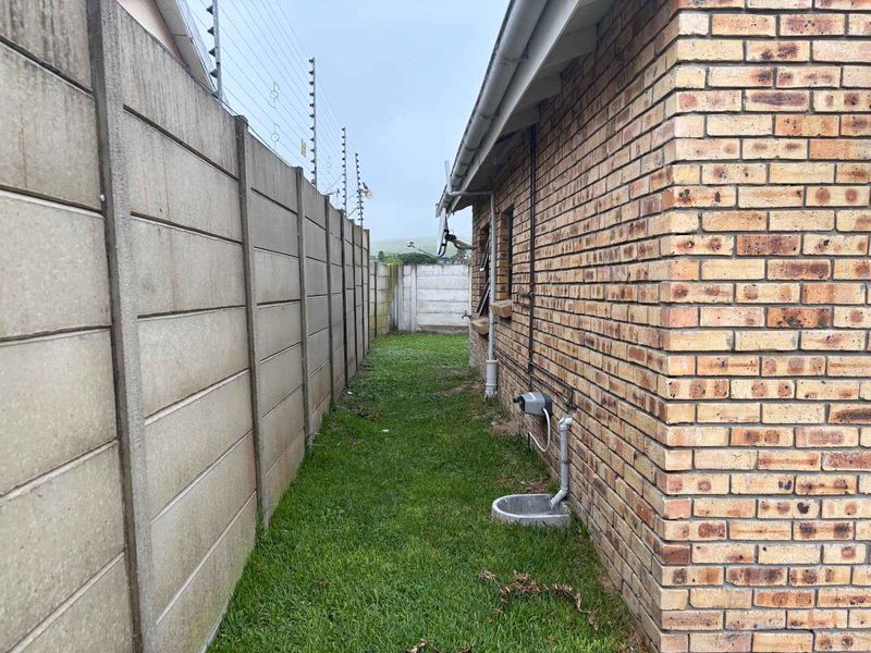 Side passage of the cottage featuring face brick exterior walls, precast concrete boundary fencing with electric security wires, and established lawn area.