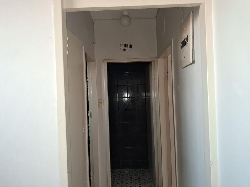 A functional apartment hallway featuring a central light fixture, ventilation grille, and an electrical distribution board, leading towards a tiled bathroom area.