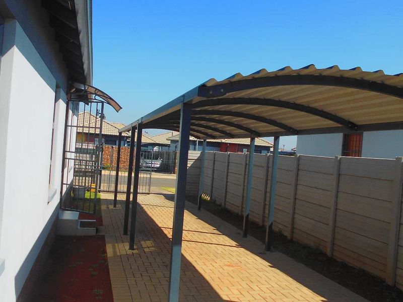 A functional outdoor carport area featuring a metal roof structure and paved driveway, enclosed by a precast concrete boundary wall.
