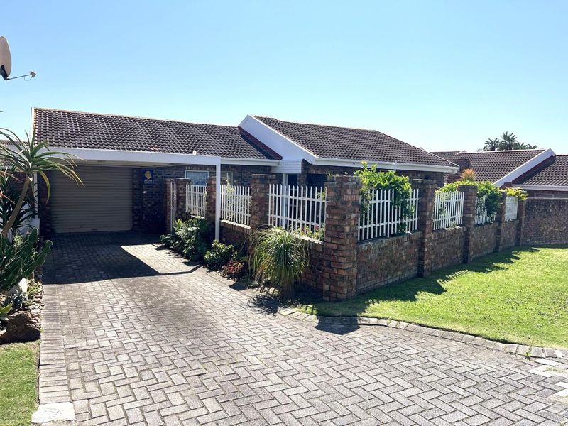 Front exterior view of a brick townhouse featuring a paved driveway, carport, and secure perimeter wall with white metal fencing.