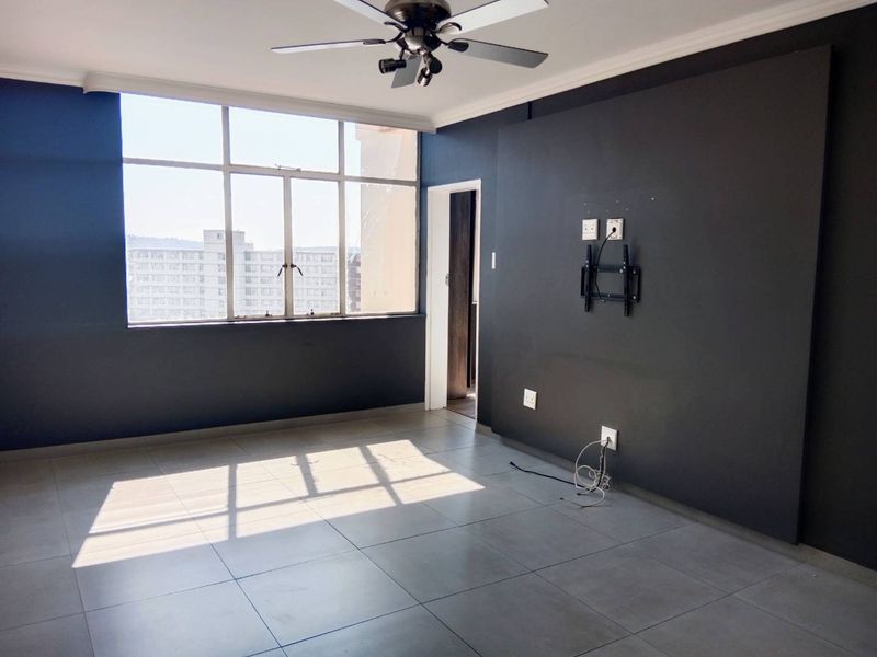 An empty living room featuring large windows, tiled flooring, a ceiling fan, and a dark accent wall with a mounted TV bracket.