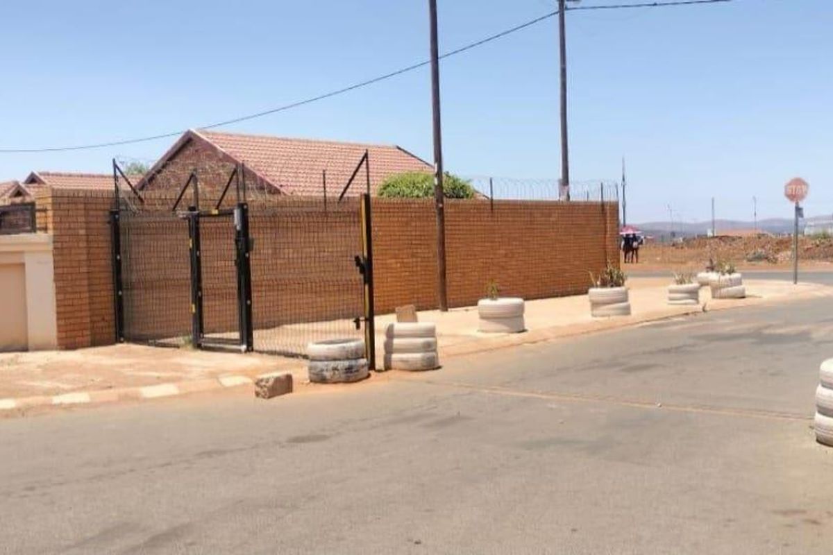 Street view of the property entrance featuring a brick boundary wall, security fencing with razor wire, and a metal gate.