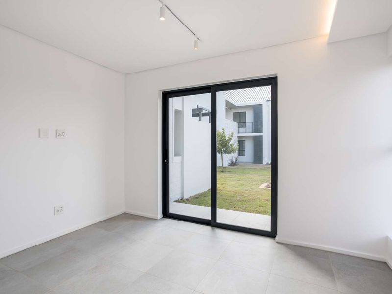 A bright, modern, and empty living area featuring large sliding glass doors that open to a private garden view, finished with neutral floor tiles.