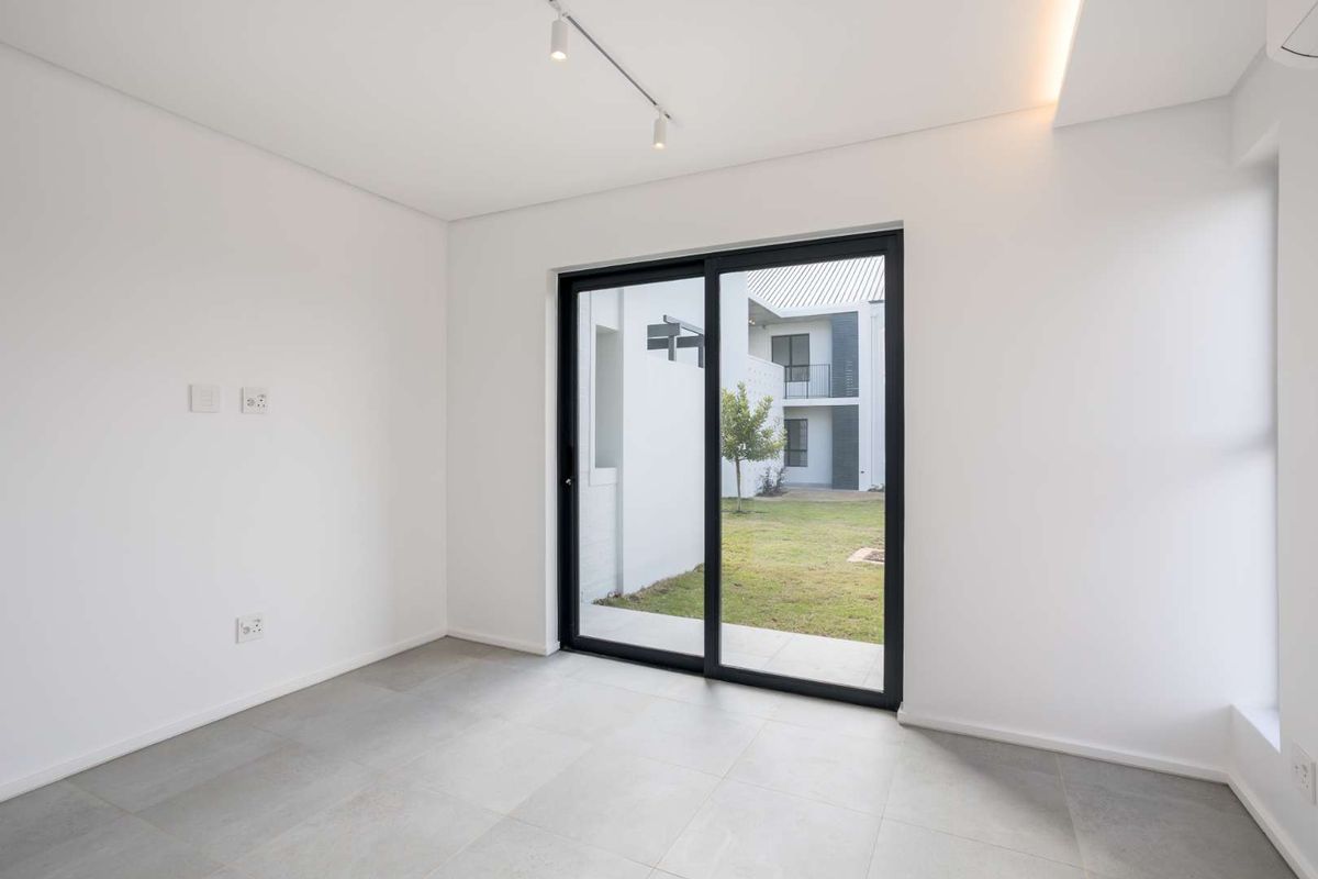 A bright, modern, and empty living area featuring large sliding glass doors that open to a private garden view, finished with neutral floor tiles.