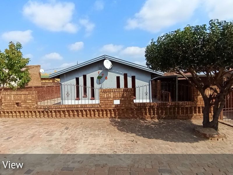 Front view of a single-story house featuring a brick boundary wall, metal security fencing, and a paved driveway area.