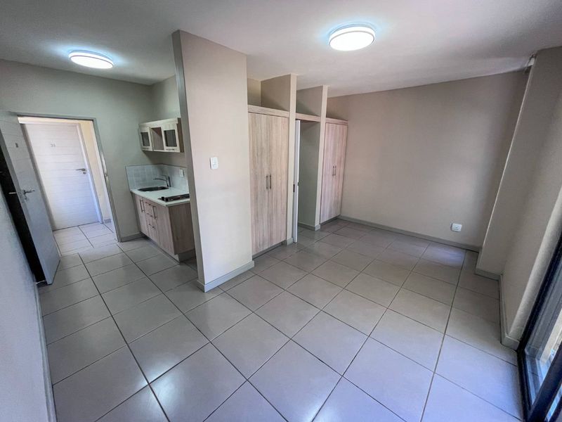 An open-plan living area featuring tiled flooring, built-in cupboards, and a compact kitchen corner with cabinetry and a sink.