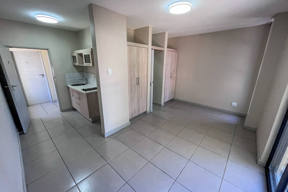 An open-plan living area featuring tiled flooring, built-in cupboards, and a compact kitchen corner with cabinetry and a sink.