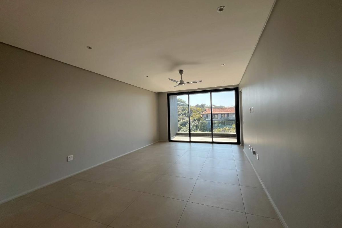 A spacious, empty living area featuring neutral tiled flooring, recessed lighting, a ceiling fan, and large sliding glass doors leading to an outdoor balcony.