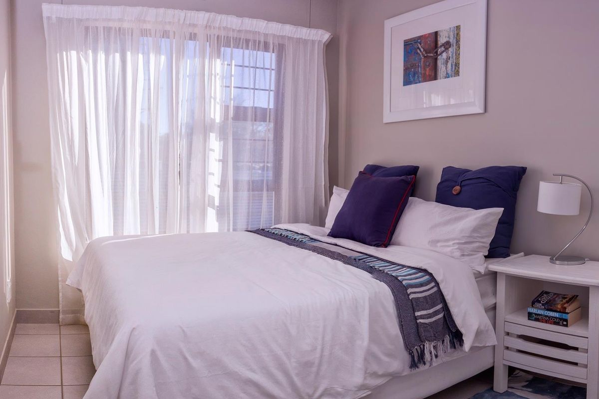 A well-lit bedroom featuring neutral wall tones, tiled flooring, and large windows with sheer curtains, creating a bright and comfortable living space.