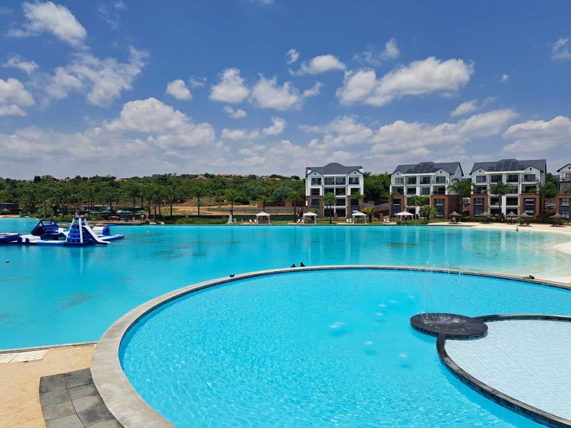 A expansive, well-maintained communal swimming pool area featuring a central water fountain and clear blue water, set within a modern apartment complex.