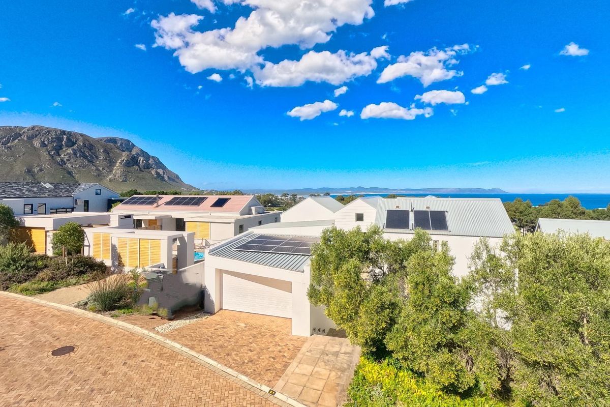 An elevated aerial view of a modern residential property featuring solar panels, a paved driveway, and a garage, set against a scenic coastal backdrop.