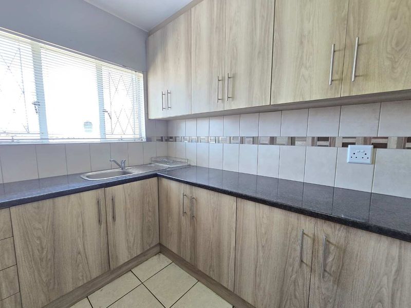 A clean and functional kitchen featuring light wood-grain cabinetry, dark granite countertops, tiled backsplash, and a stainless steel sink with natural light.
