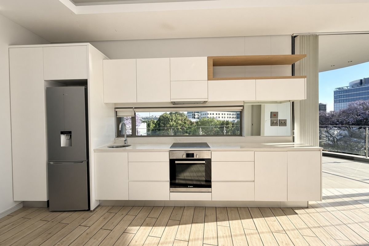 Modern, minimalist kitchen featuring sleek white cabinetry, stone countertops, integrated appliances, and wood-look tile flooring, with a large window offering natural light and views.