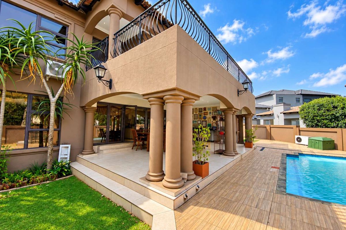 Spacious rear exterior featuring a covered patio, upper balcony with wrought iron railings, and a swimming pool set within a landscaped garden.
