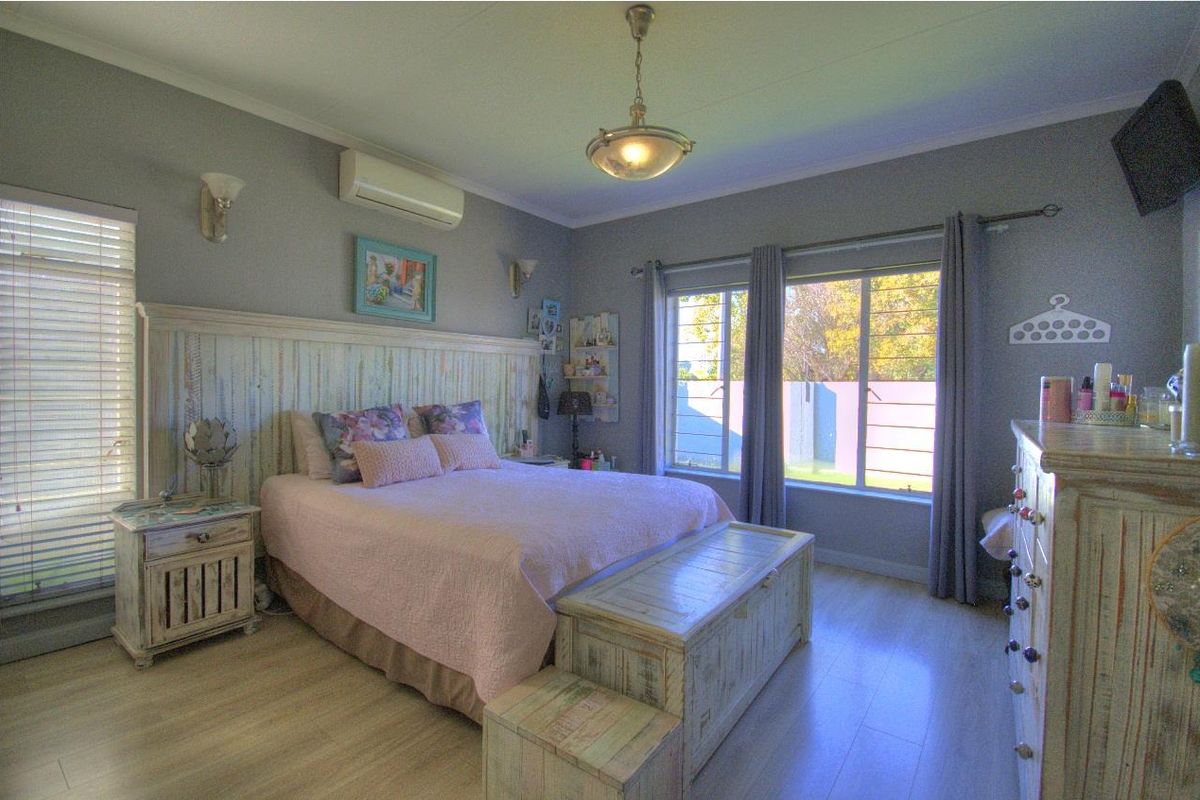A well-lit bedroom featuring light wood-look flooring, a built-in headboard, wall-mounted air conditioning unit, and large windows providing natural light.
