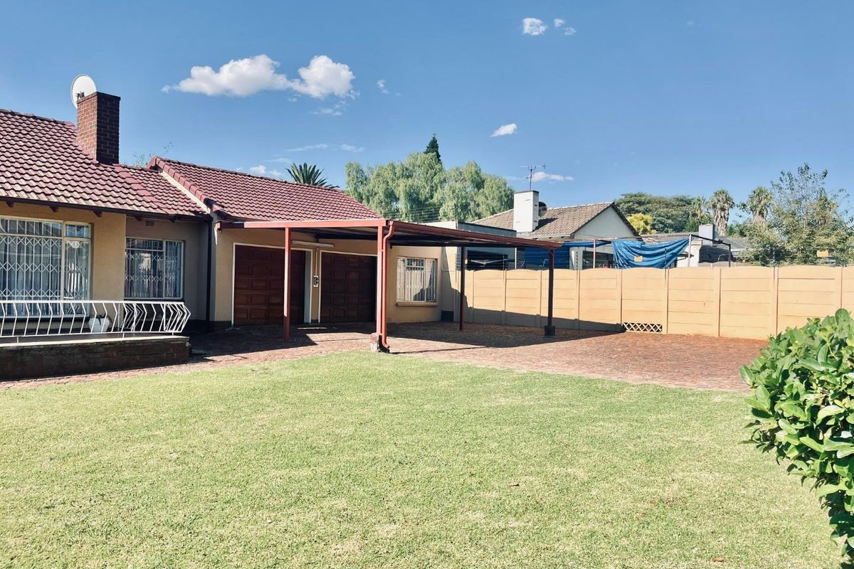 A spacious rear garden featuring a paved driveway, a covered carport, and a well-maintained lawn area.