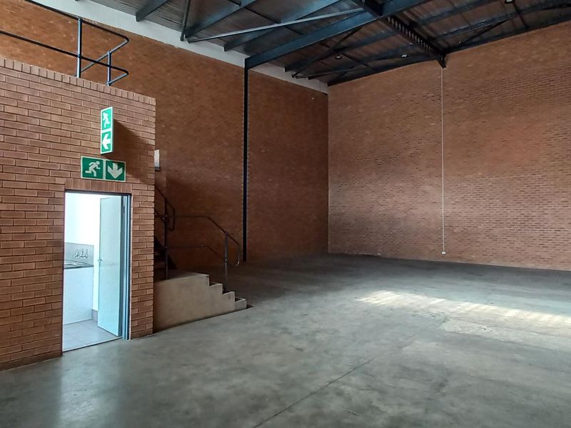Spacious industrial warehouse unit featuring high ceilings with exposed steel trusses, durable concrete flooring, and brick wall finishes with a mezzanine access point.