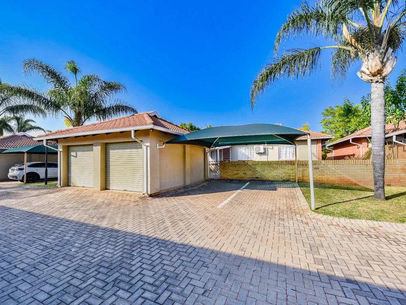 Exterior view of a townhouse featuring a double garage, an additional carport, and a paved driveway within a well-maintained residential complex.