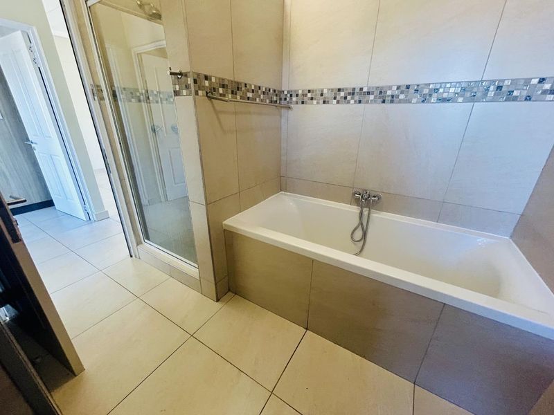 A clean, modern bathroom featuring a built-in bathtub with chrome fixtures, a separate glass-enclosed shower, and neutral floor-to-ceiling tiling with a decorative mosaic border.