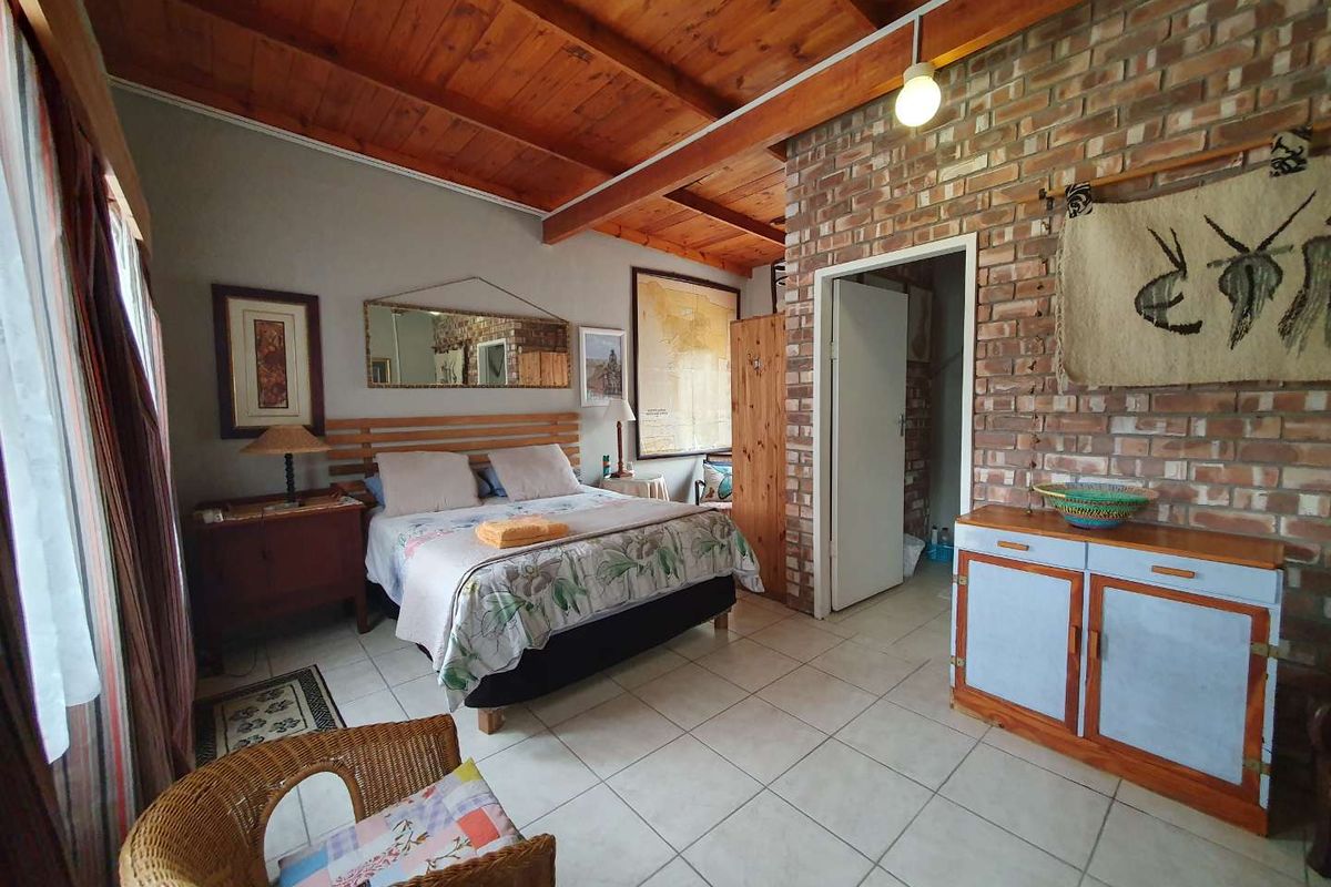 A spacious bedroom featuring exposed timber beam ceilings, tiled flooring, and a rustic brick feature wall with an adjoining doorway.