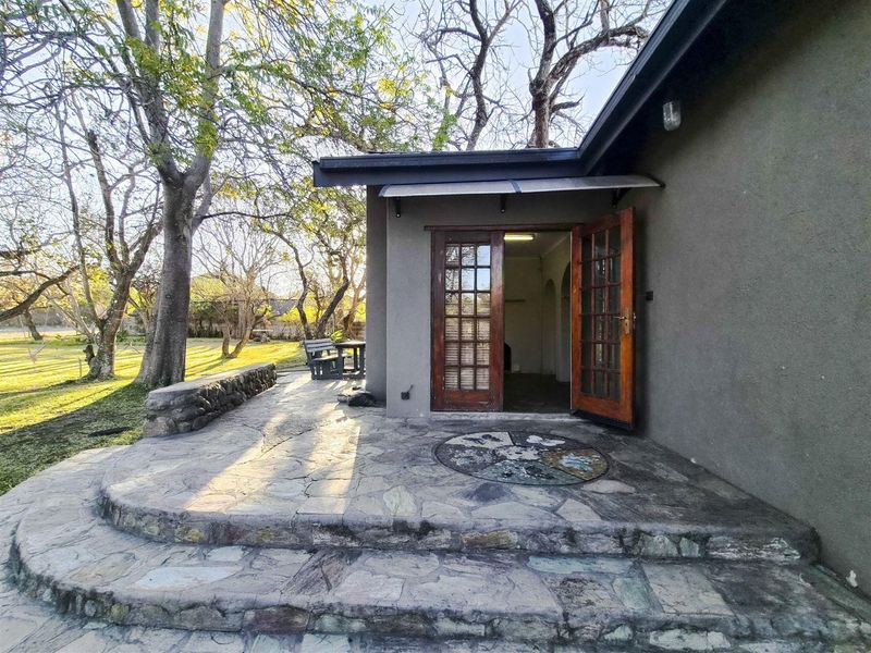 A rustic stone-paved patio area featuring a low stone retaining wall, wooden French doors, and a small protective awning, set within a garden environment.