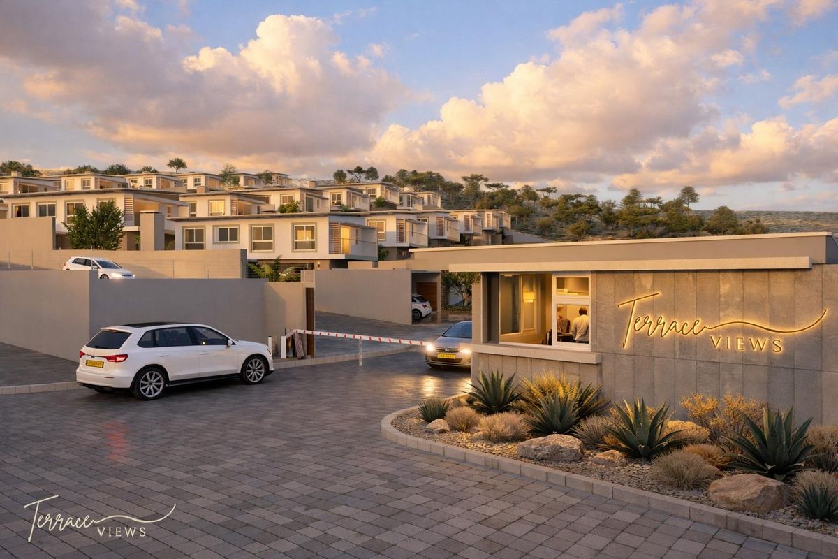 A modern residential estate entrance featuring a security gatehouse, paved driveway, and tiered townhouse architecture set against a scenic hillside backdrop.
