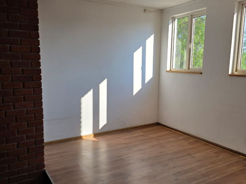 An empty, sunlit room featuring laminate flooring, an exposed brick pillar, and dual windows, offering a blank canvas for industrial or office space customization.
