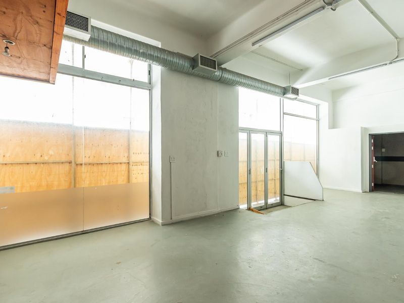 An empty, industrial-style open plan space featuring high ceilings, exposed ducting, and large windows providing ample natural light.