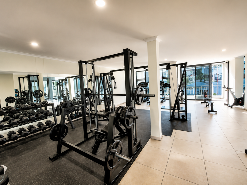 A well-maintained apartment complex gym featuring rubber flooring, mirrored walls, integrated lighting, and large sliding glass doors providing natural light.