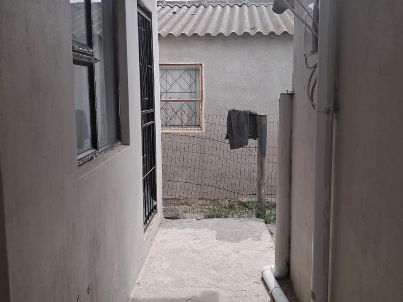 A narrow exterior side passage featuring a concrete pathway, security gate, and exposed drainage infrastructure along the building exterior.