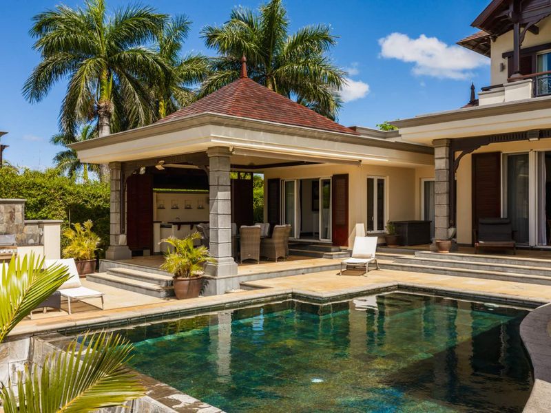 A beautifully landscaped outdoor pool area featuring a covered patio with stone columns, integrated outdoor kitchen, and direct access to the main residence.