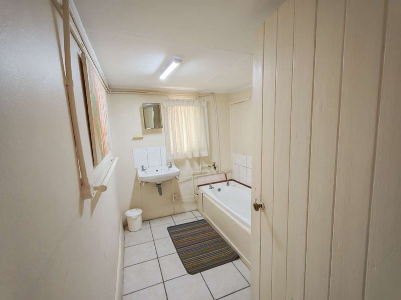 A functional bathroom featuring a bathtub, wall-mounted basin, and tiled flooring, with visible surface-mounted plumbing and dated finishes throughout.