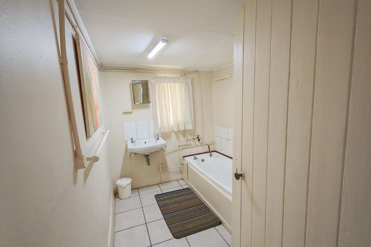 A functional bathroom featuring a bathtub, wall-mounted basin, and tiled flooring, with visible surface-mounted plumbing and dated finishes throughout.