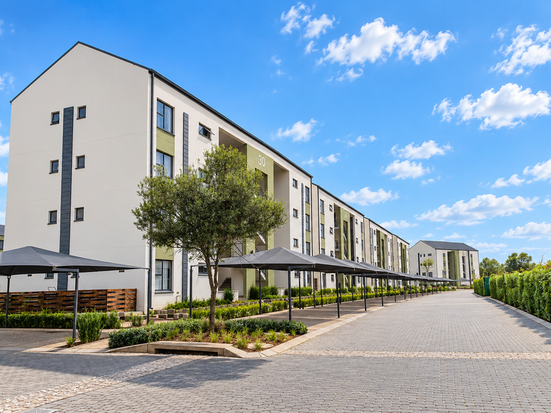 A well-maintained apartment complex exterior featuring paved driveways, covered parking structures, and neat landscaped garden beds under a clear blue sky.