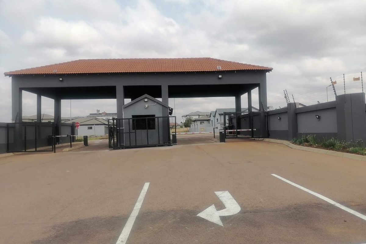 A well-maintained residential estate entrance featuring a secure gatehouse, perimeter walling with electric fencing, and a paved driveway with boom gate access.