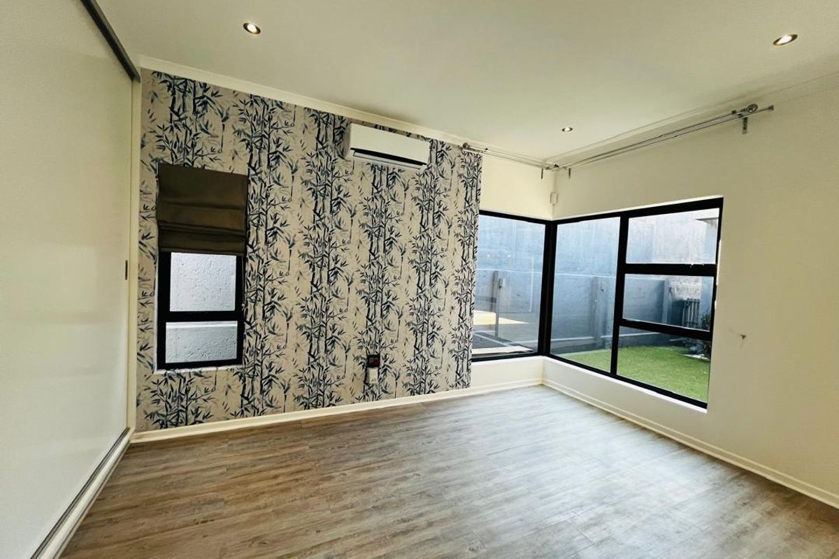 A bright, empty bedroom featuring modern laminate flooring, a built-in wardrobe, air conditioning, and large windows overlooking a private garden area.