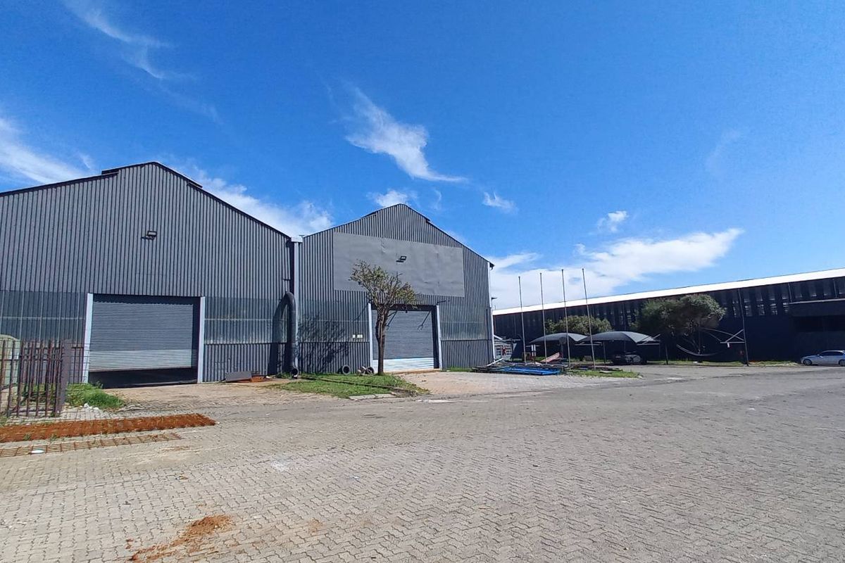 Industrial property featuring large corrugated metal warehouse structures with roller shutter doors, set within a spacious paved yard with security fencing.