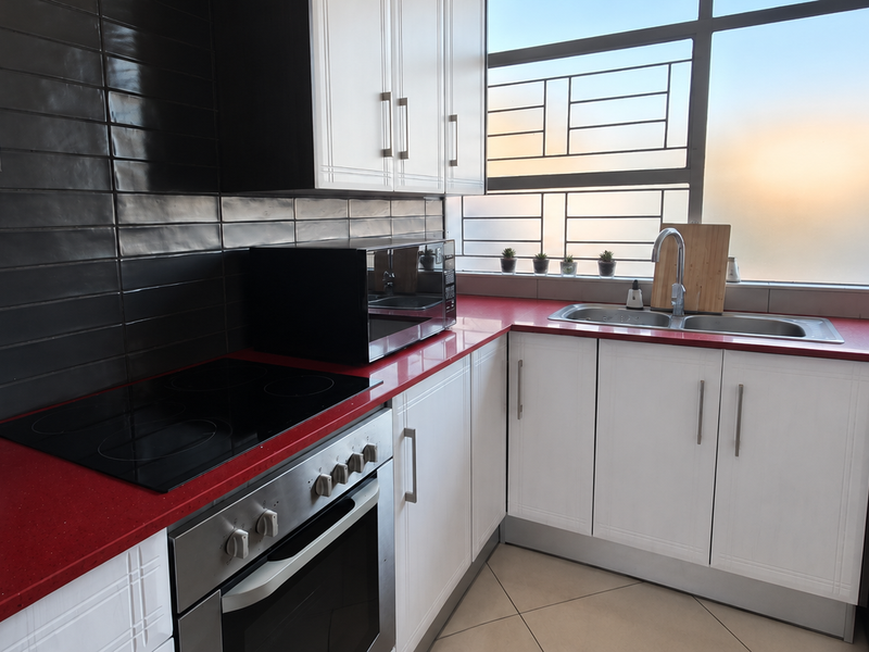 Modern kitchen featuring white cabinetry, striking red countertops, black tiled splashback, built-in oven, electric hob, and integrated washing machine with large window.
