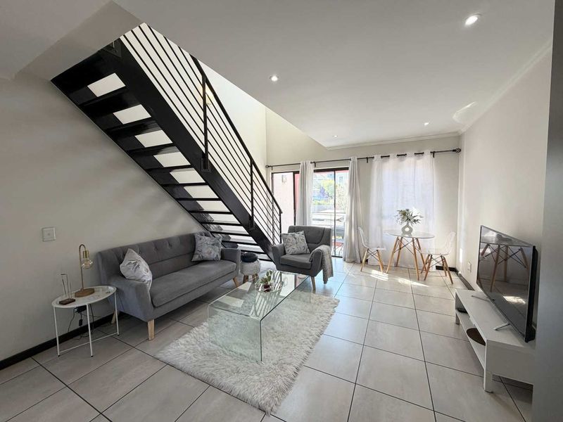 A bright, open-plan living area featuring modern tiled flooring, recessed lighting, and a striking black metal staircase leading to the upper level.