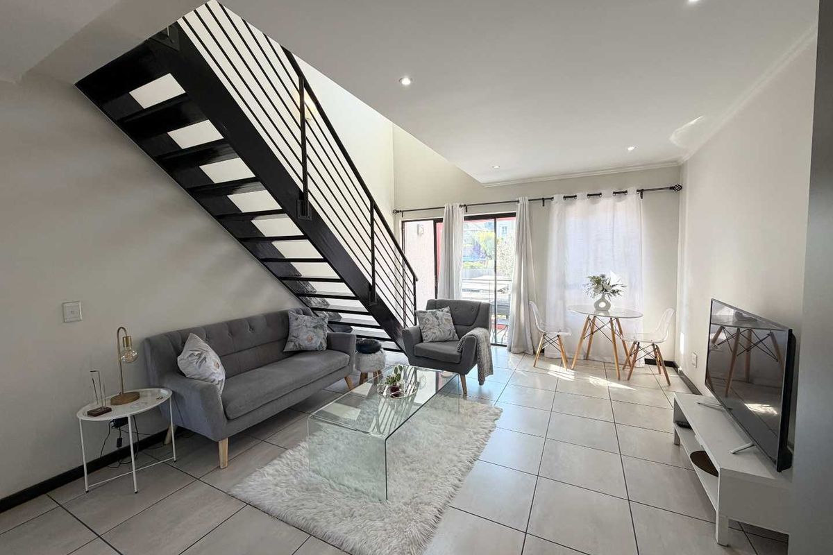 A bright, open-plan living area featuring modern tiled flooring, recessed lighting, and a striking black metal staircase leading to the upper level.