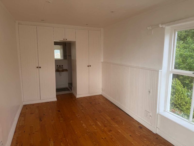 A bright bedroom featuring polished timber flooring, white wainscoting, built-in wardrobes, and a view of the garden through a large sash window.