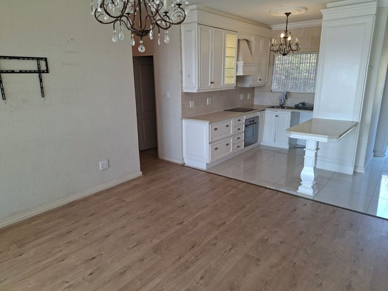 Open-plan living and kitchen area featuring white cabinetry, integrated appliances, a breakfast counter, and a combination of laminate and tiled flooring.