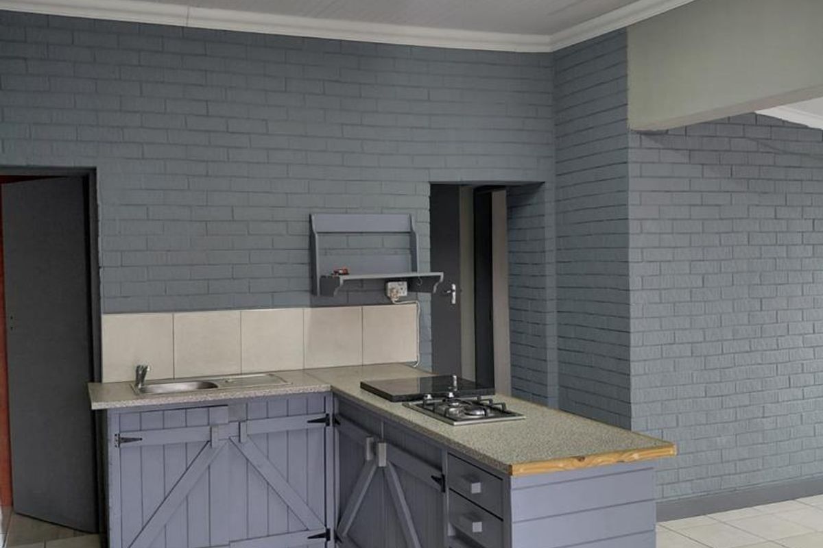 A compact kitchen featuring grey painted brick walls, custom timber cabinetry, a gas hob, and tiled flooring in an open-plan layout.