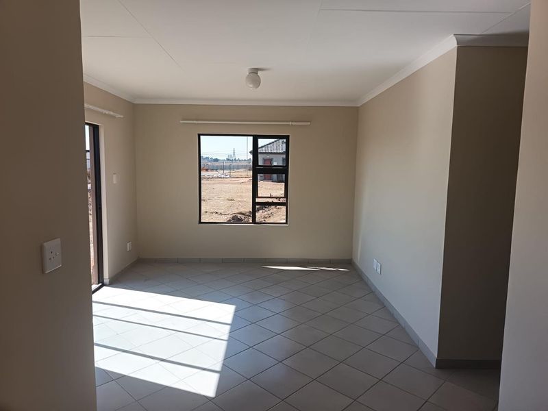 A bright, empty living room featuring neutral tiled flooring, aluminium window frames, and a sliding door leading to the exterior.