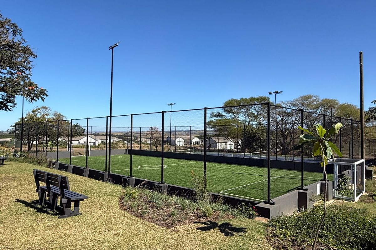 A well-maintained outdoor sports facility featuring artificial turf, perimeter fencing, and integrated floodlighting, set within a landscaped residential environment.