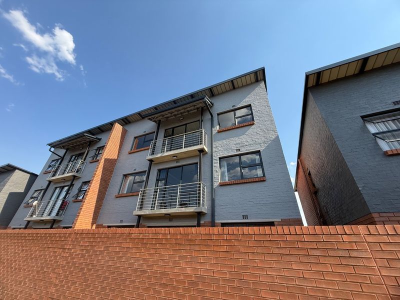 A low-angle exterior view of a modern apartment building featuring grey brickwork, multiple balconies with metal railings, and a prominent red brick boundary wall.